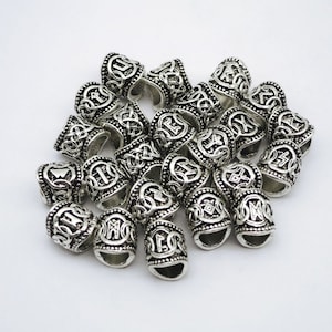 Könnte beinhalten: Ein Haufen silberfarbener Metallperlen mit komplizierten Designs. Jede Perle hat ein erhabenes Muster aus einem Knoten oder Symbol. Die Perlen sind dazu bestimmt, als Haargummis oder Haarwickel verwendet zu werden.