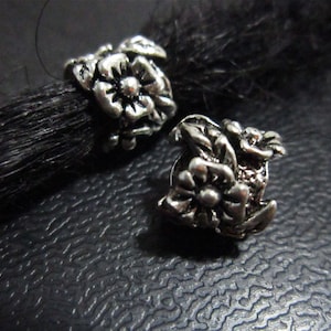 May include: Two silver metal hair beads with floral designs. The beads are in the shape of a flower with a raised center and petals.