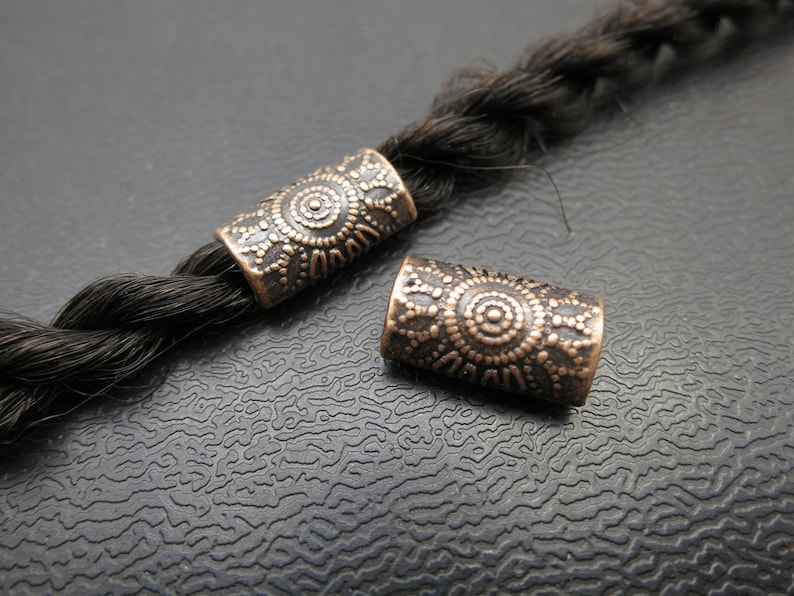 May include: Two copper hair beads with intricate circular patterns. The beads are on a dark brown braid.