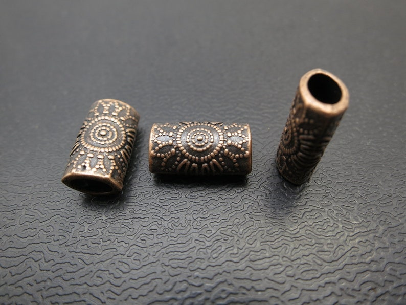 May include: Three antique copper-toned metal beads with a decorative design. The beads are cylindrical and have a circular pattern.