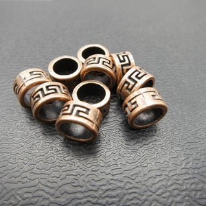 May include: A group of eight copper-colored metal beads with a geometric design. The beads are arranged in a pile on a black textured surface.