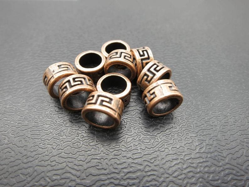 May include: A group of eight copper-colored metal beads with a geometric design. The beads are arranged in a pile on a black textured surface.