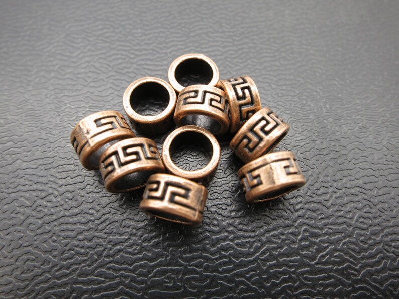 May include: A group of ten copper-colored metal beads with a black Greek key pattern. The beads are round and have a hole in the center.
