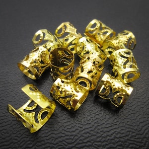 May include: A group of gold-colored hair beads with intricate designs. The beads are made of metal and have a hollow center.