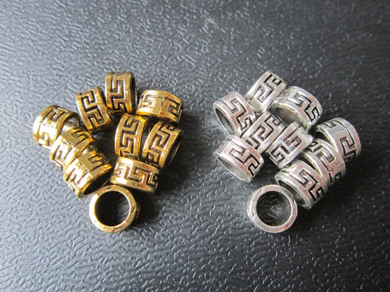 May include: A group of gold and silver metal beads with a Greek key design. The beads are stacked on top of each other and have a shiny finish.