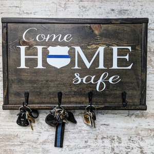 May include: Wooden key holder with the text "Come Home Safe" and a blue line police badge. There are four hooks on the bottom for hanging keys.