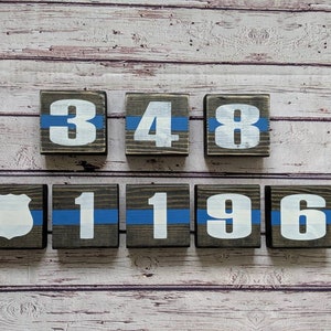 May include: Wooden blocks with white numbers and a blue line painted on each block. The numbers spell out "348 1196". There is also a wooden block with a white police badge on it.
