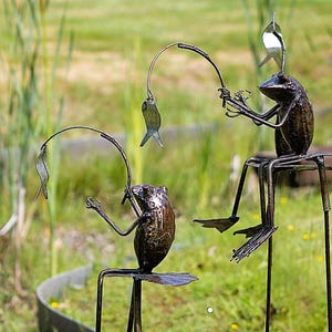 Puede incluir: Esculturas de ranas de metal pescando con cañas de alambre doblado y señuelos con forma de pez. Las ranas están sentadas en pequeños taburetes, una de ellas con una cuchara en la cabeza. El fondo presenta hierba alta.