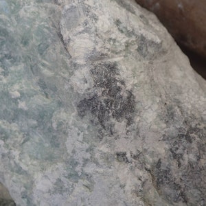 Peut inclure: Gros plan sur une pierre vert clair et grise avec un motif gris foncé. La pierre a une surface rugueuse et texturée.