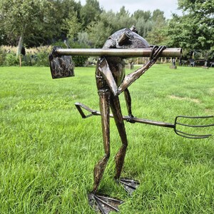 Peut inclure: Une sculpture en métal brun foncé représentant une grenouille debout sur des pattes palmées. La figurine tient une pelle à long manche sur ses épaules et porte une fourche en métal dans une main, avec une finition texturée et patinée.