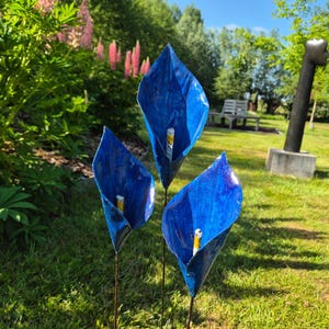Könnte beinhalten: Drei leuchtend blaue Metallblumenskulpturen mit gelben Zentren stehen in einem grasbewachsenen Garten. Die Gartenkunst ist vor dem Hintergrund von grünem Laub, rosa Blumen und einem sonnigen Himmel platziert. Die Skulpturen stehen auf dünnen Metallstielen.