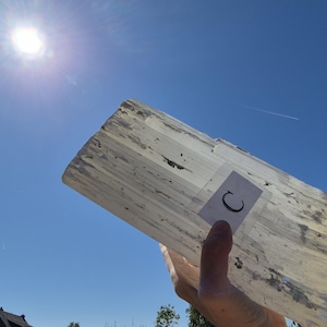 Op de afbeelding: Een rechthoekige plaat van doorschijnend wit selenietkristal met een vezelige, gelaagde textuur en zichtbare minerale insluitingen. Een kleine witte sticker met de zwarte letter C is op het oppervlak van de steen bevestigd tegen een blauwe lucht.