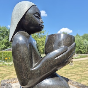 May include: A dark stone sculpture of a person holding a bowl. The figure is kneeling, with a calm expression. The sculpture has a smooth, reflective surface. The background shows greenery and a blue sky with clouds.