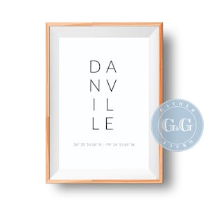 Puede incluir: Una impresión enmarcada con la palabra "DANVILLE" en una fuente minimalista. La impresión está en un marco de madera clara e incluye las coordenadas 36° 35 14.06" N -79° 24 15.85" W. Un logotipo circular azul y blanco está en la esquina inferior derecha.