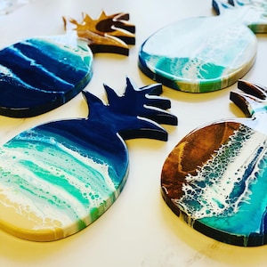 May include: Five resin pineapple coasters with a blue, white, and brown ocean wave design. The coasters are round and have a raised pineapple design.