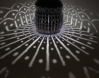 May include: A black metal lantern with a decorative design casts a patterned shadow on a dark surface. The shadow features a repeating pattern of white lines and dots.
