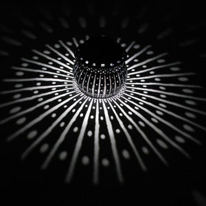 May include: A black and white patterned lantern casts a geometric shadow on a dark surface. The lantern has a round shape and a textured surface.