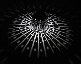 May include: A black and white patterned lantern casts a geometric shadow on a dark surface. The lantern has a round shape and a textured surface.