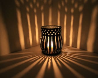 May include: A black metal candle holder with a decorative pattern casts intricate shadows on a white surface. The candle holder is lit, creating a warm glow.