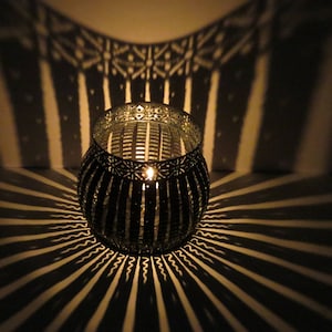 May include: A black metal candle holder with a geometric design casts a patterned shadow on a white surface. The candle holder has a round shape and is lit with a candle inside.