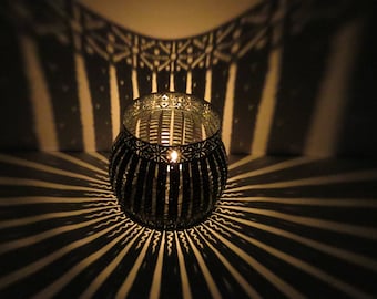 May include: A black metal candle holder with a geometric design casts a patterned shadow on a white surface. The candle holder has a round shape and is lit with a candle inside.