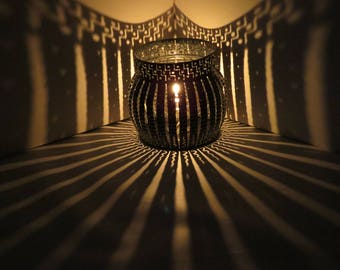May include: A black metal candle holder with a geometric design casts intricate shadows on a white surface. The candle holder has a round shape and is lit by a single candle.