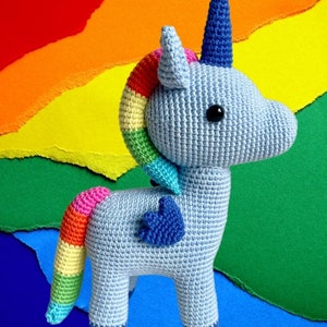 May include: A crocheted blue unicorn with a rainbow mane and tail. The unicorn has a blue horn and a heart-shaped wing. The unicorn is standing on a colorful background.