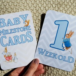 Może przedstawiać: Dwie karty kamieni milowych dla dziecka z niebieskim tekstem i ilustracjami. Jedna karta głosi "Baby Milestone Cards" z ilustracją wiewiórki. Druga karta głosi "1 Week Old" z ilustracją królika.