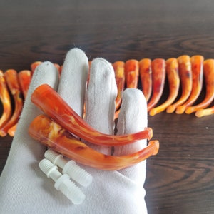 Könnte beinhalten: Zwei orangefarbene und weiße marmorierte Pfeifenköpfe mit weißen Kunststoffverbindern. Die Köpfe sind gebogen und haben eine glänzende Oberfläche. Die Köpfe werden in einer behandschuhten Hand gehalten.