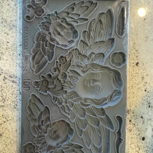 May include: A gray silicone mold featuring cherub faces with detailed wings and floral accents. The package reads "IRON ORCHID DESIGNS" and "CLASSICAL CHERUBS IOD". The mold includes various decorative elements for crafting and design projects.