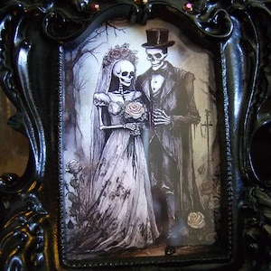 May include: A black ornate picture frame with a gothic wedding theme. The frame features a couple of skeletons dressed in wedding attire, holding a bouquet of flowers. The frame is decorated with rhinestones.