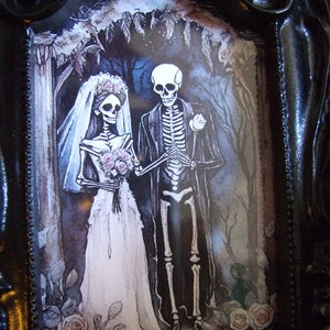 May include: A black ornate picture frame with a white mat and a printed image of a skeleton bride and groom in a forest setting. The bride is wearing a white gown and veil, and the groom is wearing a black suit. The image is surrounded by a black frame with ornate details.
