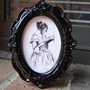 May include: Black oval picture frame with a detailed, ornate design. The frame surrounds a black and white illustration of a woman reading a book. The frame has decorative jewels at the corners.