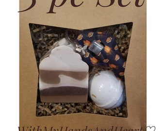 May include: A 3-piece gift set in a brown box, including a bar of soap, a bath bomb, and a small accessory. The soap is light brown and white, the bath bomb is white, and the accessory is silver. The box has the text "3 pc Set" and "WithMyHandsAndHeart".