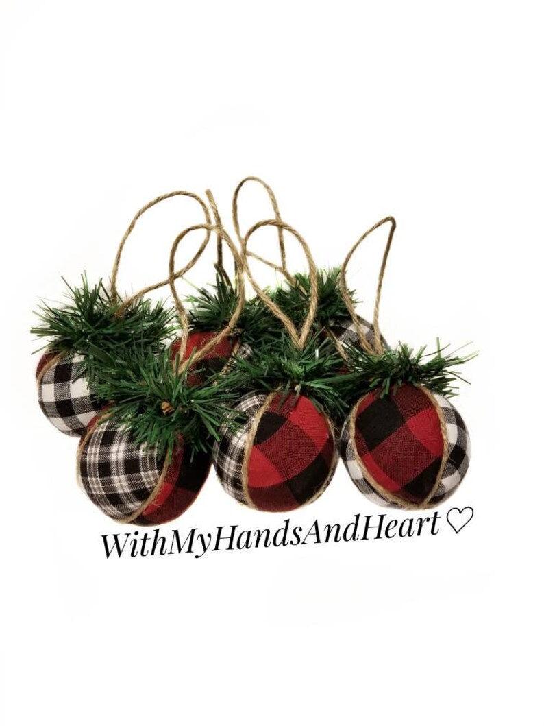 May include: A set of five spherical ornaments covered in red, black, and white buffalo plaid fabric. Each ornament features a small cluster of green artificial pine needles at the top and a brown twine loop for hanging. The text WithMyHandsAndHeart is visible.