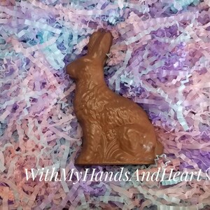 May include: A molded milk chocolate rabbit figurine in a seated position, featuring textured fur details and floral accents at the base. The text WithMyHandsAndHeart is printed in white at the bottom center of the image over shredded paper.