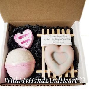 May include: A gift box containing a heart-shaped soap dish, a heart-shaped soap, and a bath bomb. The soap dish is made of wood and the bath bomb is pink and white. The box is filled with black paper shreds. Text on the box reads "WithMyHandsAndHeart".
