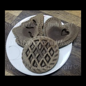 May include: Three brown, textured, circular and heart-shaped baked goods with embossed patterns, including a lattice design, a small heart cutout, and a leaf-like shape, arranged on a white plate with the text WithMyHandsAndHeart.