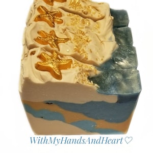 May include: A rectangular bar of soap featuring a beige top layer decorated with four gold starfish and gold glitter accents. The side of the soap displays a marbled pattern of beige, blue, and gold swirls. The text WithMyHandsAndHeart is below.