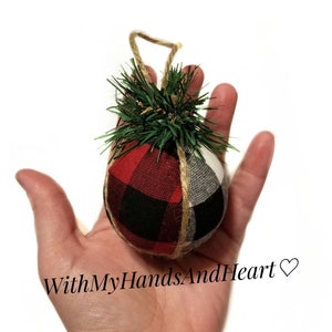 May include: A round fabric ornament featuring a red and black buffalo plaid pattern on one side and a white and black buffalo plaid pattern on the other. The ornament is topped with green artificial pine needles and a jute twine hanging loop.
