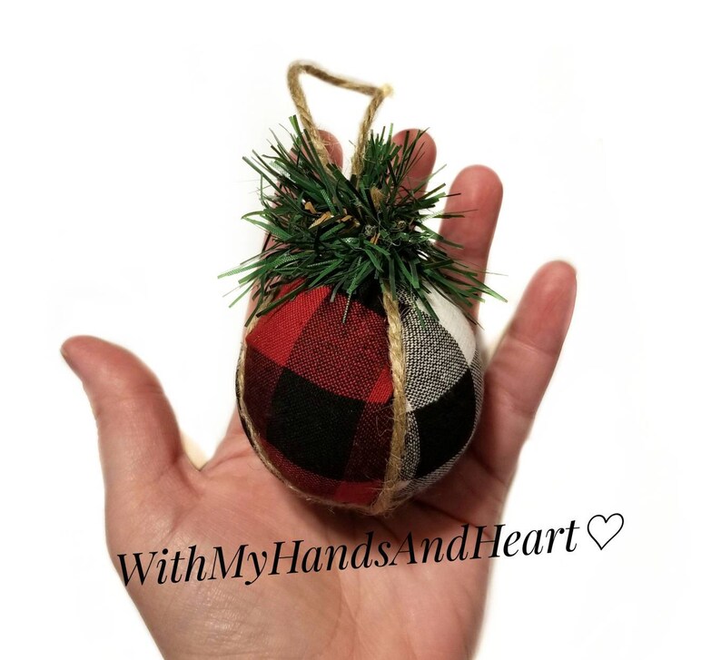 May include: A round fabric ornament featuring a red and black buffalo plaid pattern on one side and a white and black buffalo plaid pattern on the other. The ornament is topped with green artificial pine needles and a jute twine hanging loop.