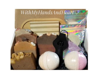 May include: A gift box labeled WithMyHandsAndHeart containing three rectangular soap bars with textured tops, two round bath bombs, a black keychain holder, and two iridescent pouches filled with small bath products on a black paper filler.
