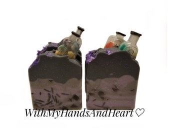 May include: Two rectangular bars of dark purple and grey soap, each topped with embedded miniature glass bottles containing colored liquids. The soap features a layered color gradient and dark, jagged inclusions throughout the base. Text reads WithMyHandsAndHeart.