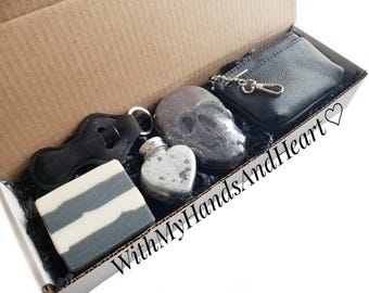 May include: A gift box containing a black and white striped rectangular soap bar, a heart-shaped glass bottle with a silver cap, a dark gray skull-shaped object, and a black zippered pouch with a silver keychain. The text WithMyHandsAndHeart♡ is visible.