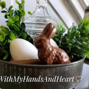 May include: A rustic metal container holds a chocolate bunny, white eggs, and green foliage. A clear glass object and a lamp are in the background. The text "WithMyHandsAndHeart" is at the bottom of the image.
