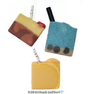 May include: Three handmade soap bars designed to resemble beverages. One is brown, tan, and yellow with a cherry and a straw. Another is blue with dark brown circles and a black straw. The third is yellow with a straw.
