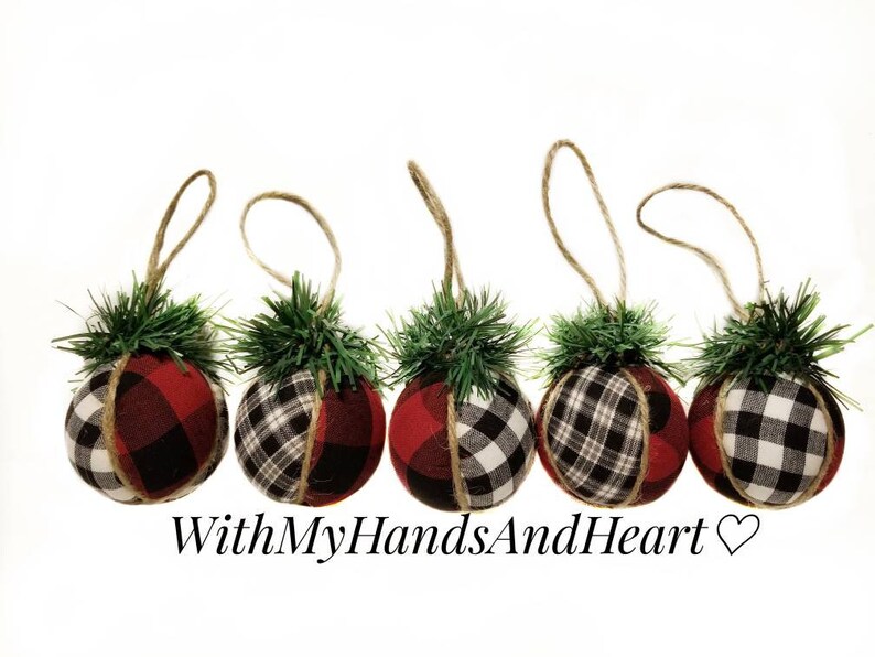 May include: Five spherical ornaments wrapped in red and black buffalo plaid fabric, each topped with a small cluster of green pine needles and a jute twine hanging loop. The text WithMyHandsAndHeart is printed below the ornaments.