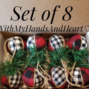 May include: A set of 8 fabric-covered spherical ornaments in a cardboard box labeled "SET of 8" and "WithMyHandsAndHeart". Each ornament features a red, black, and white buffalo plaid pattern, accented with green pine needles and jute twine.