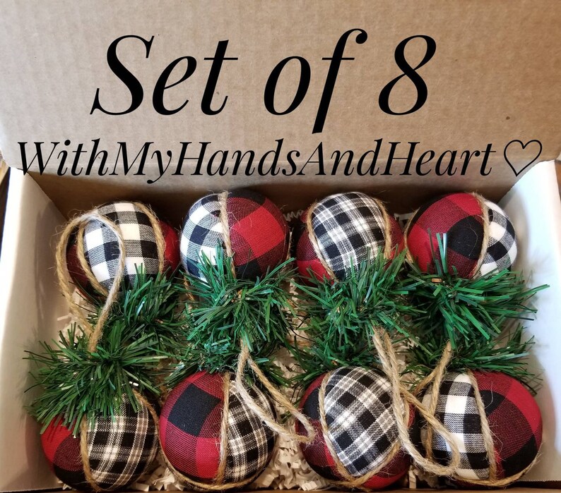 May include: A set of 8 fabric-covered spherical ornaments in a cardboard box labeled "SET of 8" and "WithMyHandsAndHeart". Each ornament features a red, black, and white buffalo plaid pattern, accented with green pine needles and jute twine.