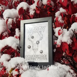 May include: A framed black and white print with the text "MERRY CREEPMAS!" features a deer skull with ornaments. The print is set against a backdrop of red leaves covered in snow, creating a festive, yet spooky holiday theme.
